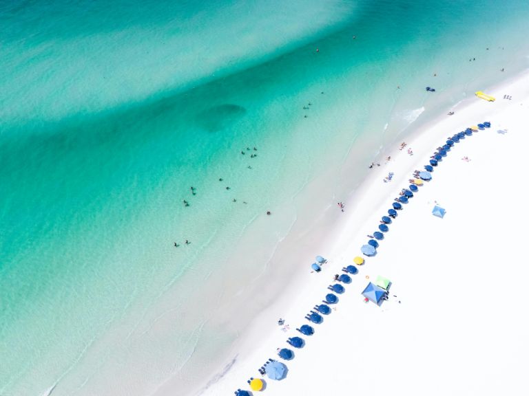 Emerald Coast waters meet a sugar-white sand beach with blue umbrellas and chairs for visitors.