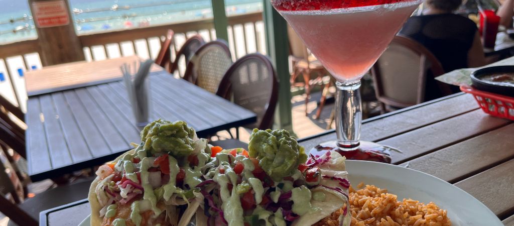 Fresh fish tacos and a pink margarita sit on a high-top table on a deck overlooking the Emerald Coast of Florida.