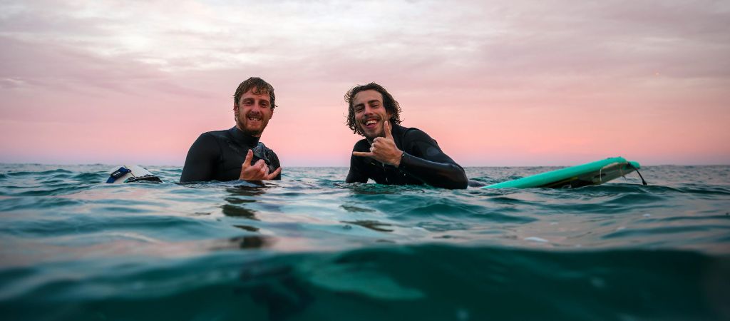 Two young men in wet suits wave a hang loose hand sign while sitting on their surf boards in the ocean, with a pink and purple sunrise in the background.