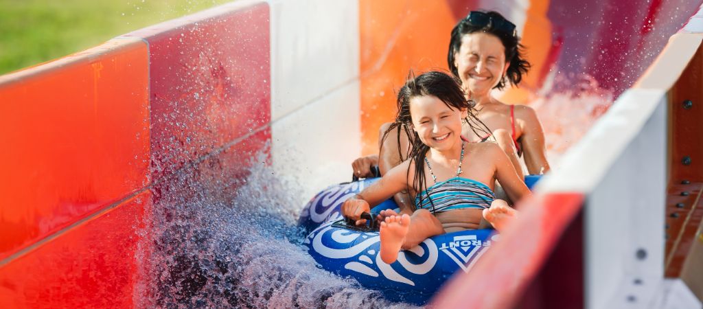 A smiling mother and young daughter ride a double inflatable tube down a colorful waterslide.