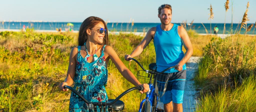 A smiling woman and man walk their bicycles from the beach along a sandy path, with tall beachgrass all around and the coast in the background.