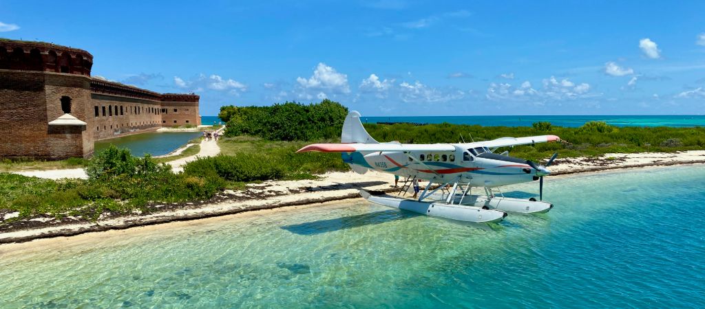 A seaplane rests at the coastal water's edge in front of Fort Jefferson on a sunny day.