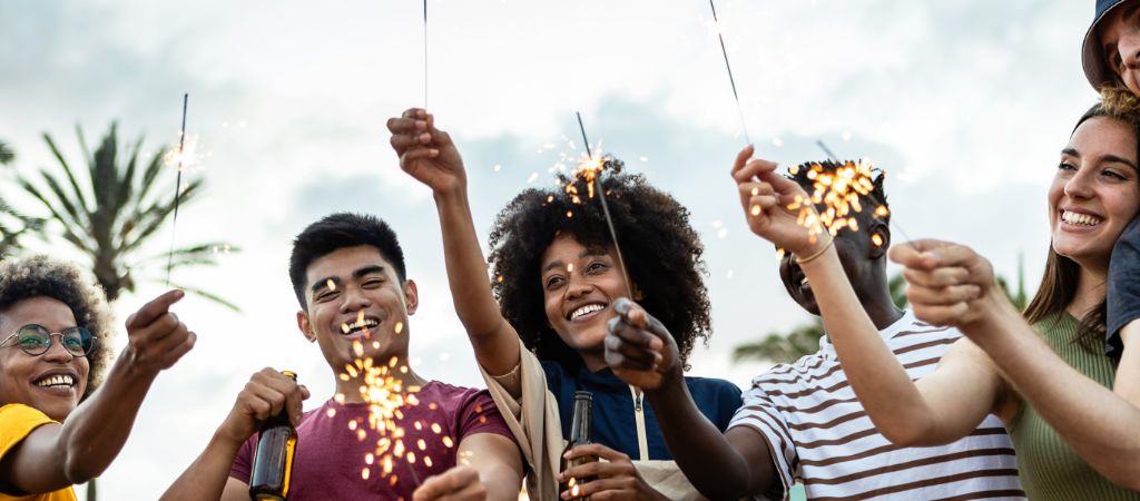 Group of young people with sparklers and glass-bottled beer smiling on a beach with palm trees in the background.