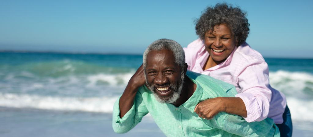 An older woman climbs onto the back of an older man, both grinning ear to ear, on a white-sand beach in the Florida Panhandle, with lapping waves in the background.