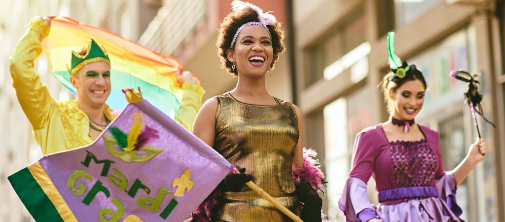 A group of 3 people dressed in colorful attire and makeup stand on a float in a downtown parade waving a Mardi Gras flag.