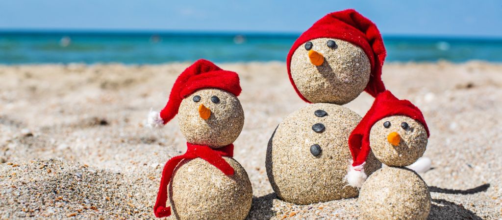 Three small snowmen made of sand wear red Christmas hats on a beach with the Emerald Coast in the background.