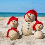 Three small snowmen made of sand wear red Christmas hats on a beach with the Emerald Coast in the background.