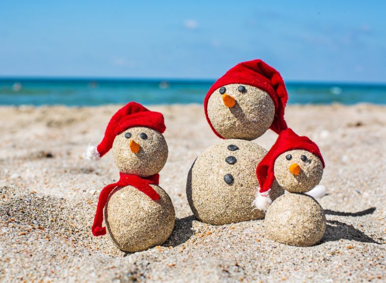 Three small snowmen made of sand wear red Christmas hats on a beach with the Emerald Coast in the background.