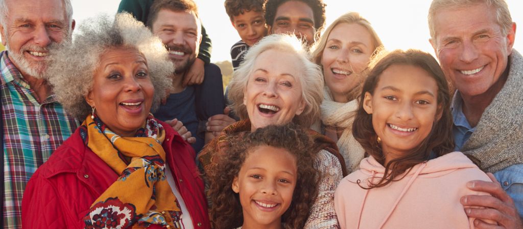 Group of 11 family members, old to young, grinning ear to ear in light sweater weather on a beach.