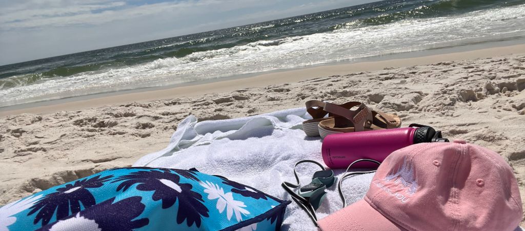 A white towel with a beach bag, hat, water bottle, sandles, and goggles sits on Perdido Key Beach on a sunny day, with waves in the background.