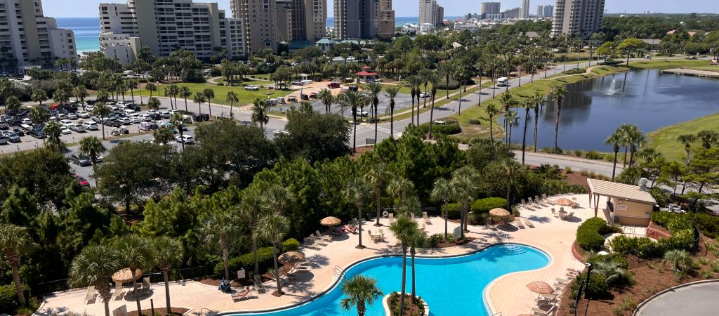 Overview of Sandestin Golf & Beach Resort from a high-rise condo balcony, with a resort pool, pond, towering buildings, and distant ocean views.