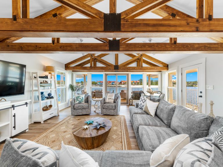 A photo of the inside of a bright beach house with wood beams over a living area, looking out to ocean views through floor-to-ceiling windows.