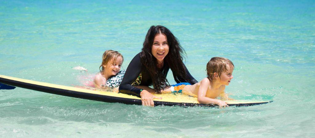 Mother and two small children smile while floating on a boggie board on a sandbar.