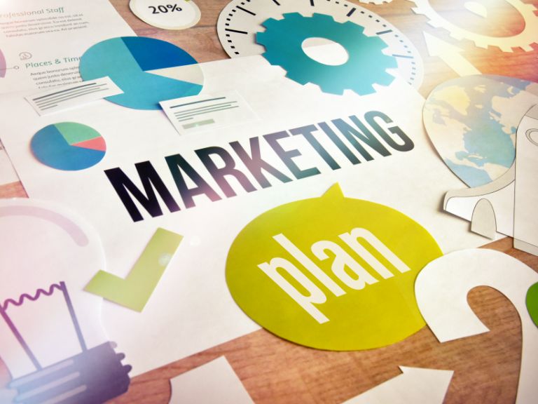 Overhead shot of desk with reference materials and marketing symbols that says "Marketing Plan."