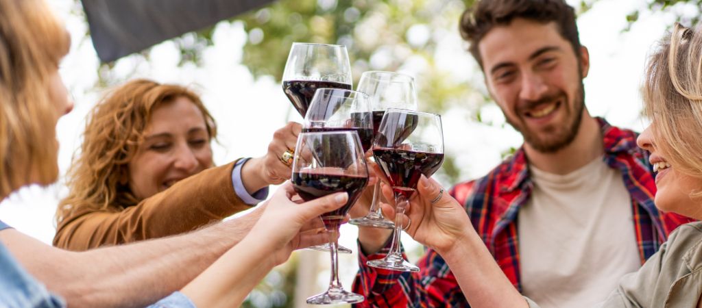 A group of people toast glasses of wine while smiling at each other beneath an outdoor tent on a sunny day.