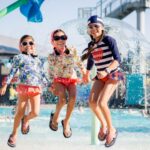 Three girls jump on a splash pad at a waterpark on a sunny day, smiling at the camera.