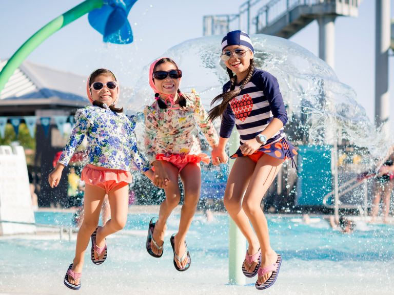 Three girls jump on a splash pad at a waterpark on a sunny day, smiling at the camera.